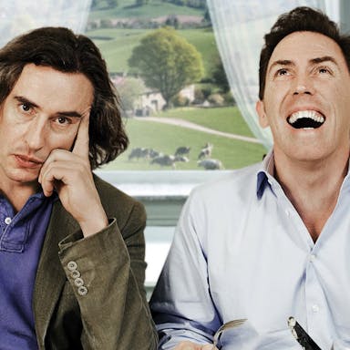 Two middle aged white men sitting at a table with the countryside behind them in the window, one laughing, one looking fed up 