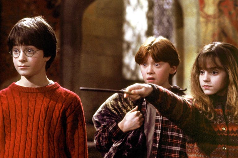 Two white boys and a white girl look surprised at something. One of the boys is wearing a red jumper, the other a check shirt. The girl is wearing a jumper and is pointing a wand somewhere off screen.