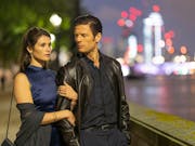 A woman holds on to a man's arm as they stand by a river in a city.