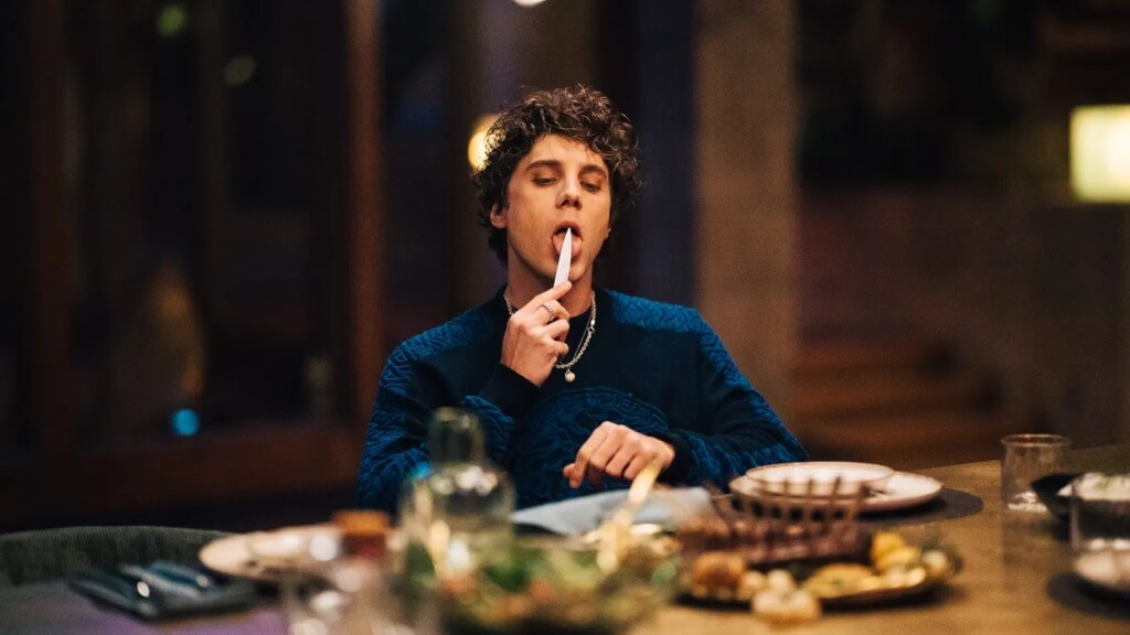 A young white boy sitting at a large dinner table, holding a knife up to his mouth and licking it