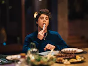 A young white boy sitting at a large dinner table, holding a knife up to his mouth and licking it