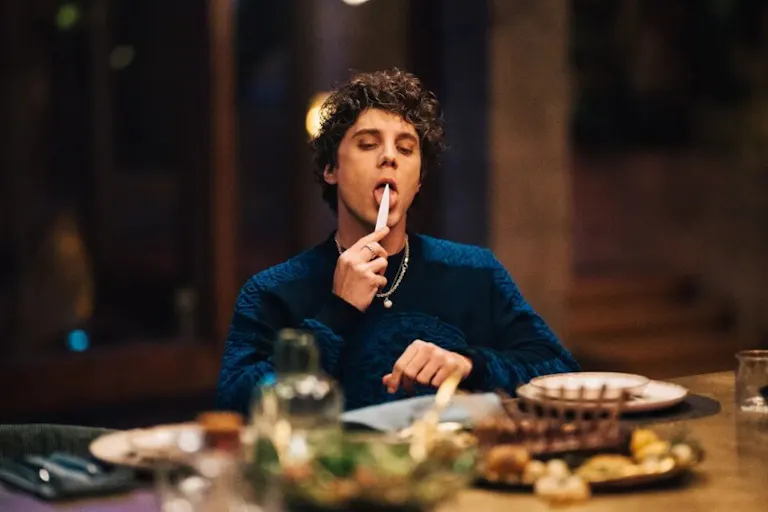 A young white boy sitting at a large dinner table, holding a knife up to his mouth and licking it