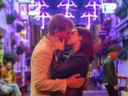 A white man and white woman kissing in a vibrant neon lit side street with bars and restaurants