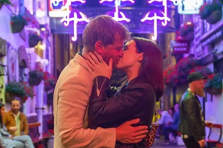 A white man and white woman kissing in a vibrant neon lit side street with bars and restaurants 