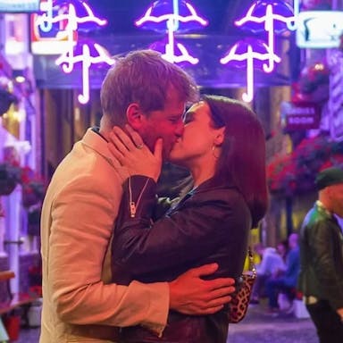 A white man and white woman kissing in a vibrant neon lit side street with bars and restaurants 