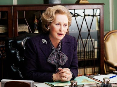 A comanding middle aged white woman with quaffed blonde hair sits at a desk 