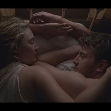Dark image of a white male and female couple laying in bed on their front, arms around each other 
