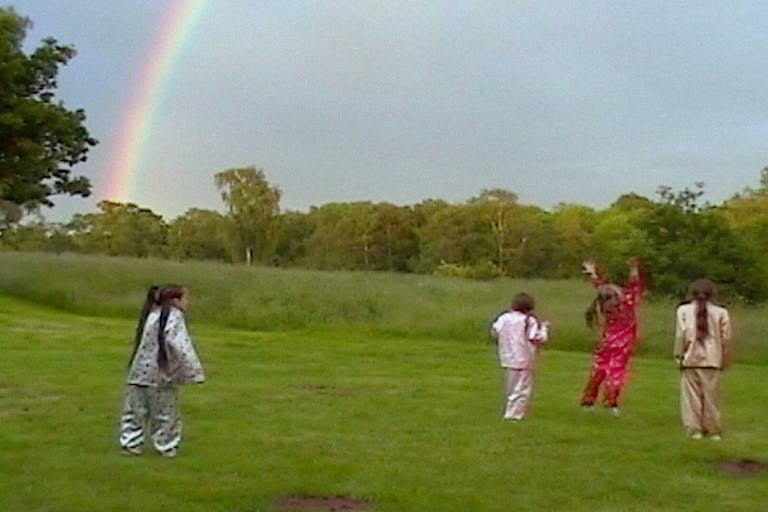 Four young white girls in shiny pajamas running about a field with a rainbow in the background