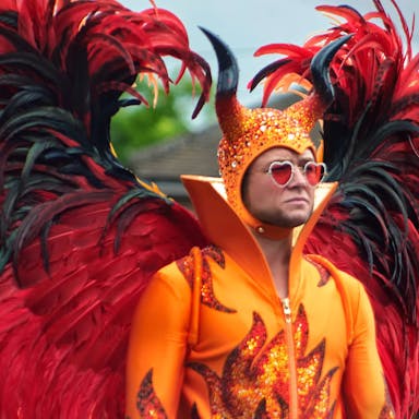 A man in red heart sunglasses and an extravagant costume, orange flame bodysuit and massive red and black feathered wings 