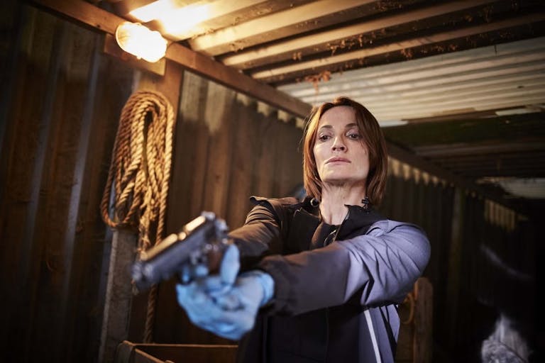 A woman in a shipping container, wearing blue forensic gloves, pointing a gun low