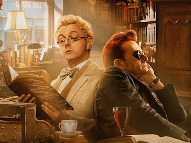 A blond haired man reading a leather bound book sits back to back with a dark red haired man with dark, steampunk glasses