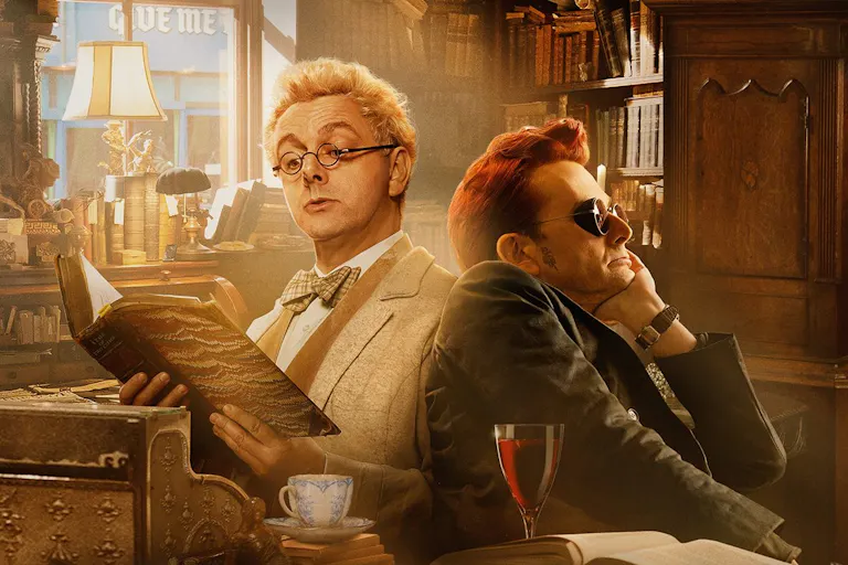 A blond haired man reading a leather bound book sits back to back with a dark red haired man with dark, steampunk glasses