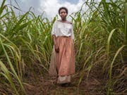 A Black woman with cropped hair, wearing a loose blouse and skirt and peach apron, walking through a plantation of tall crops looking determined