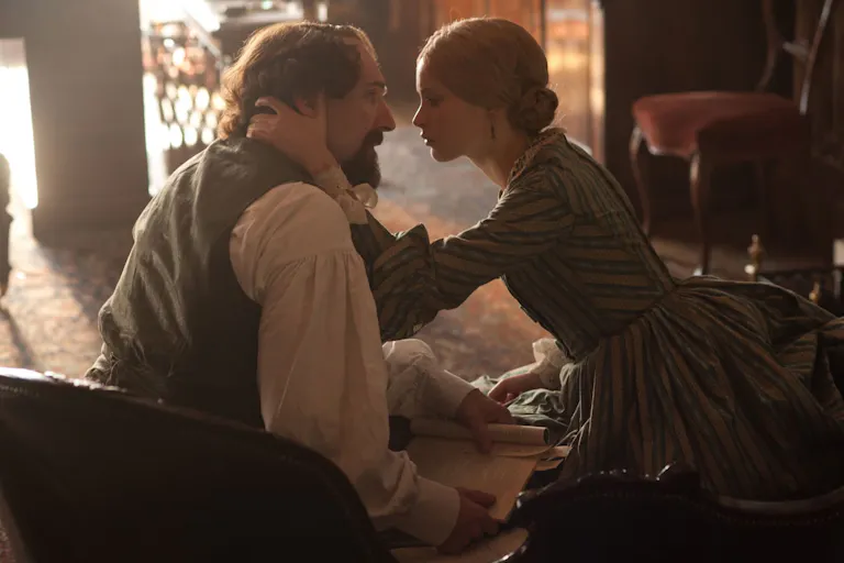 A young white woman holds the head of an older white man with long hair and beard, staring intently into his eyes, wearing period clothing