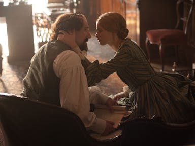 A young white woman holds the head of an older white man with long hair and beard, staring intently into his eyes, wearing period clothing