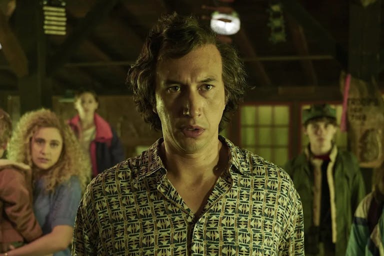 Worried looking man with short dark hair stands in the foreground, bathed in yellow light, a blonde woman holding a small child in the background.