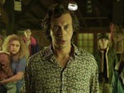 Worried looking man with short dark hair stands in the foreground, bathed in yellow light, a blonde woman holding a small child in the background.