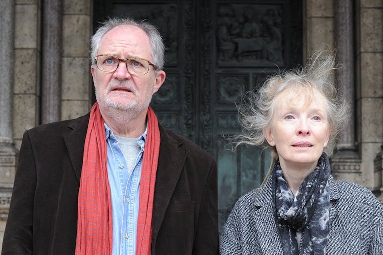 A man and a woman, both with grey hair, stand outside of an imposing looking stone building