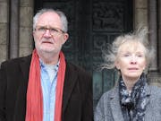 A man and a woman, both with grey hair, stand outside of an imposing looking stone building