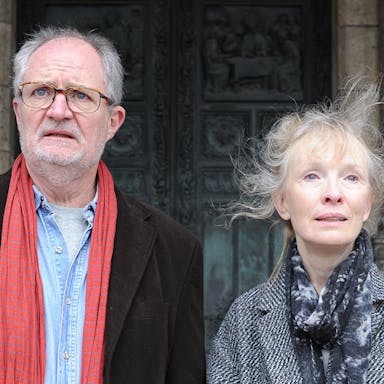 A man and a woman, both with grey hair, stand outside of an imposing looking stone building