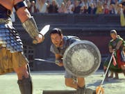 A man in gladiator armour and silver shield with sword lunges at another man in gladiator amour in a stadium of onlookers