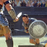 A man in gladiator armour and silver shield with sword lunges at another man in gladiator amour in a stadium of onlookers