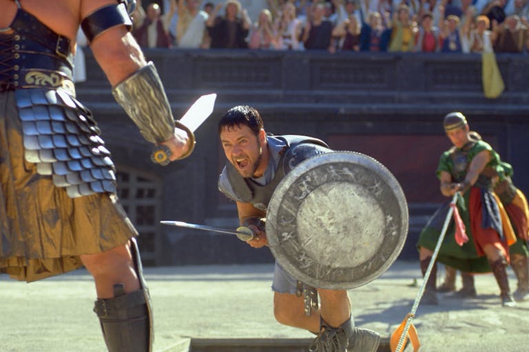 A man in gladiator armour and silver shield with sword lunges at another man in gladiator amour in a stadium of onlookers