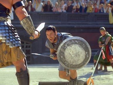 A man in gladiator armour and silver shield with sword lunges at another man in gladiator amour in a stadium of onlookers