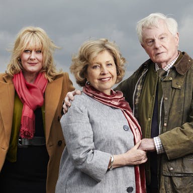 Two older white women, white older man and white middle aged woman standing in a grey countryside wearing thick coats