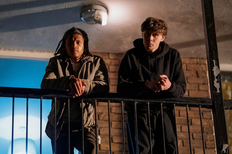 Two young men leaning over a balcony in a high rise looking down, at night