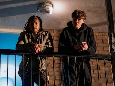 Two young men leaning over a balcony in a high rise looking down, at night