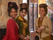 A young Black woman, brown woman and white woman smiling in a grand house wearing 1800s elegant clothing