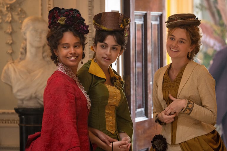 A young Black woman, brown woman and white woman smiling in a grand house wearing 1800s elegant clothing