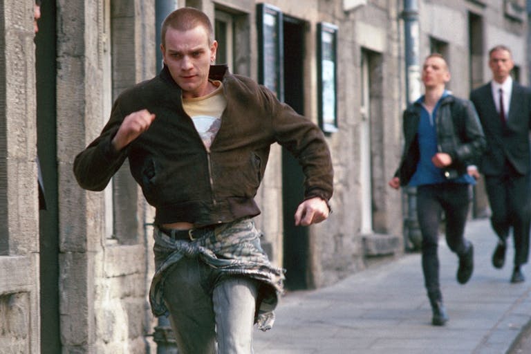 A young white man with shaved head running through a street with a another white man struggling to keep up behind him