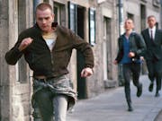 A young white man with shaved head running through a street with a another white man struggling to keep up behind him