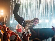 A young white man in black jeans, white boots and a tiger print jacket plays at a piano as he floats in the air