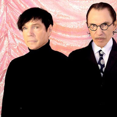 Two men in black outfits stand against a pink sparkly fabric backdrop.
