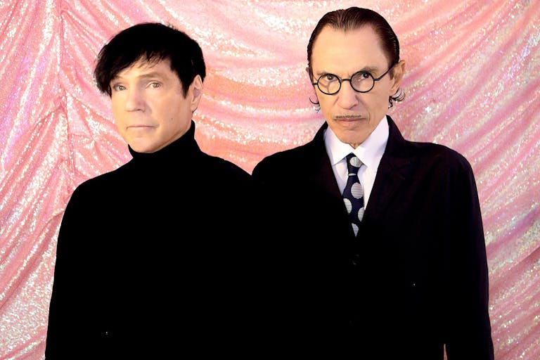Two men in black outfits stand against a pink sparkly fabric backdrop.