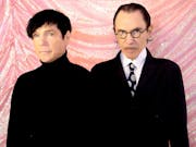 Two men in black outfits stand against a pink sparkly fabric backdrop.