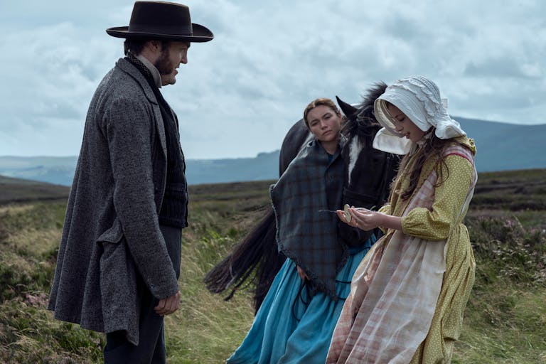 Two women and a man in old-fashioned clothes stand in the countryside outside, next to a black horse