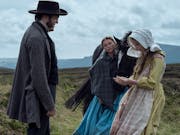 Two women and a man in old-fashioned clothes stand in the countryside outside, next to a black horse