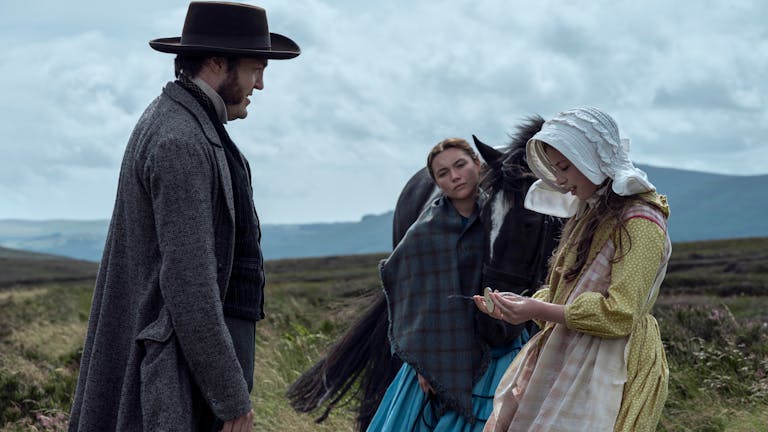 Two women and a man in old-fashioned clothes stand in the countryside outside, next to a black horse