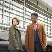 A white woman and back man in a stylish suit and coat, stand by a car and glass fronted offices