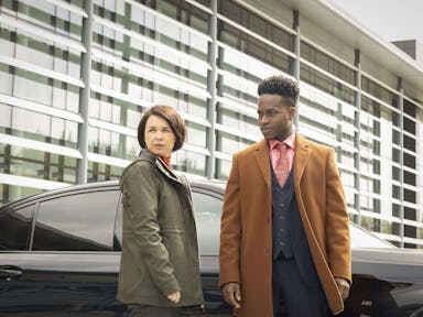 A white woman and back man in a stylish suit and coat, stand by a car and glass fronted offices