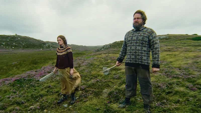 A man and woman standing on a grassy hill, looking threatening as they look ahead holding a weapon in one hand