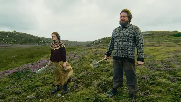 A man and woman standing on a grassy hill, looking threatening as they look ahead holding a weapon in one hand