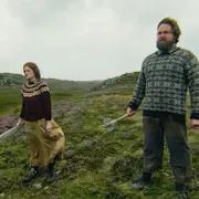 A man and woman standing on a grassy hill, looking threatening as they look ahead holding a weapon in one hand