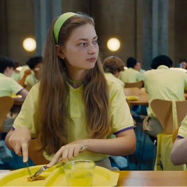 Two young white girls in yellow school uniforms staring at each other as they cut into a small, single piece of food on their yellow plastic lunch plate