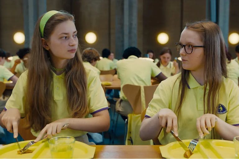Two young white girls in yellow school uniforms staring at each other as they cut into a small, single piece of food on their yellow plastic lunch plate