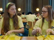Two young white girls in yellow school uniforms staring at each other as they cut into a small, single piece of food on their yellow plastic lunch plate